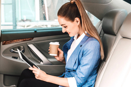 女实业家工作与数字平板电脑和在豪华轿车后座捧杯咖啡