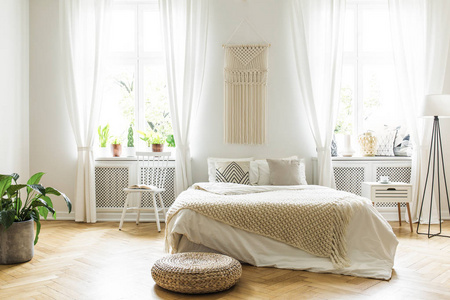 脚凳旁边的床与毯子在白色卧室内部与植物和窗户。真实照片