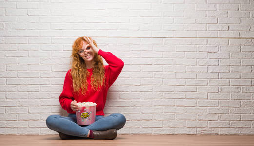 年轻红头发的妇女坐在砖墙上吃爆米花与愉快的面孔微笑做 ok 标志用手在眼睛看通过手指