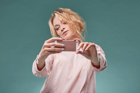 年轻的金发女郎，波浪形的头发，在光条粉红色的裙子诱人看粉色智能手机在工作室里，青绿色背景上的肖像