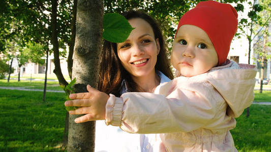 快乐的母亲在城市公园里抱着一个孩子。婴儿在日落时在树上玩树叶