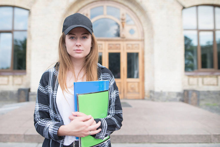 在大学建筑背景下, 穿着休闲服装的学生女孩的肖像, 看着相机。严肃的学生站立与书和笔记在校园里