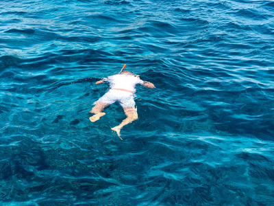 一个穿着白色 t恤短裤和面具的男性游泳者, 带着呼吸管的水肺潜水眼镜漂浮在海水的条纹上, 大海带着他的脸, 看着海底。