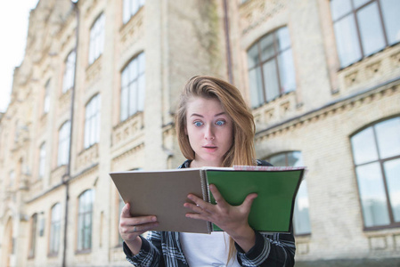 有趣的情绪化的女生看着一本书, 惊讶地看着大学大楼的背景。一个学生的肖像, 看着他的手中的书惊讶