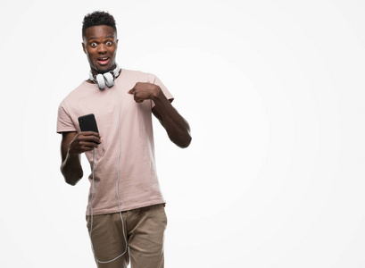 年轻的非洲裔美国男子戴着耳机, 手持智能手机与惊讶的脸指向自己的手指