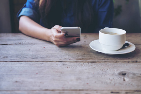 一个女人手持和使用智能手机的感觉放松, 而坐在老式咖啡馆与咖啡杯在木桌上