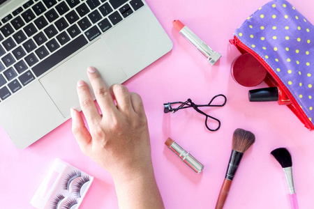 女性手的顶部视图使用笔记本电脑与化妆袋和化妆品设置在粉红色的背景。平躺