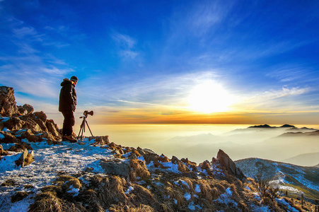 专业摄影师带照片在岩石峰上三脚架上的相机在日落时分