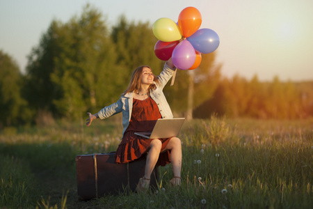 幸福的年轻女人坐在大手提箱拿着五颜六色的气球