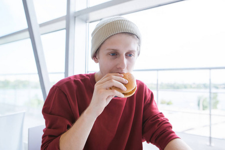 一个时尚的年轻人的肖像, 在他手中的汉堡包在一个光的背景。学生坐在一间清淡的快餐店里, 咬着一个汉堡包。快餐概念