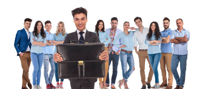 年轻的商人集团领导为您提供他的手提箱, 而他的休闲团队是站在后面的白色背景