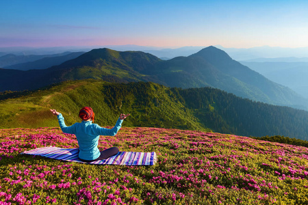 五颜六色的地毯。在莲花姿势的瑜伽女孩。草地上有杜鹃花花。高山。神奇的森林。冥想。放松。夏天的风景。温暖的早晨太阳光线