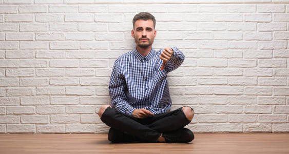 年轻的成年男子坐在白色砖墙看着不高兴和愤怒显示拒绝和否定与拇指向下手势。错误的表达式