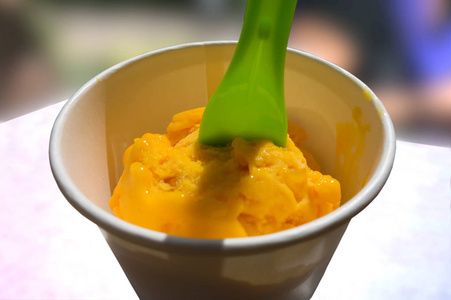 明亮的黄色的冰淇淋球在一个白色的纸杯与绿色勺子在白色和蓝色的背景。清洁饮食, 健康, 素食, 减肥, 碱性饮食食品概念