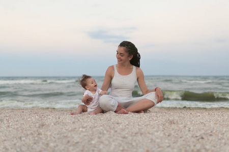 快乐的家庭母亲和孩子的女儿做瑜伽, 冥想在莲花在海滩上的位置日落