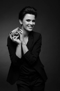年轻人微笑女人摄影师与黑色短发穿着黑色复古西装外套摆在黑暗的工作室，控股旧照片相机在她手，在黑色和白色