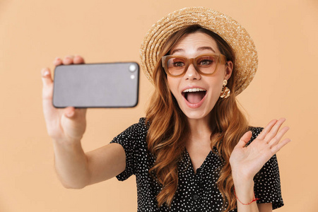 内容的画像现代妇女戴着草帽和太阳镜笑着, 并采取自拍照片的智能手机在米色背景下隔离