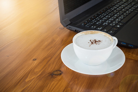 艺术在木制的桌子与计算机笔记本电脑背景上的白色杯拿铁咖啡