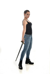 穿黑色单和牛仔裤的黑发女孩的全长肖像。站立姿势, 拿着一把剑。在白色工作室背景下被隔离