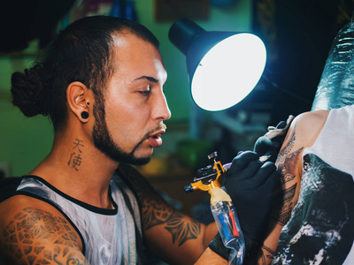 人纹身大师与长发绺显示创作纹身过程对女性的身体，在灯下光的肖像。专业的艺术家在沙龙，工作关闭视图