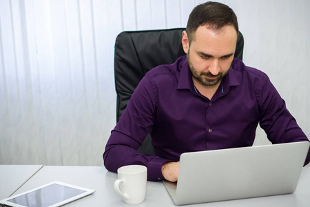 穿着紫色衬衣的男人正坐在办公室里用笔记本电脑工作。在桌上一块药片和一杯