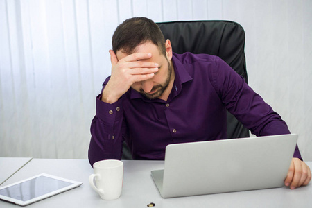 一个穿着紫色衬衣的担心的男人坐在办公室里, 抱着他的头。桌上有一块药片, 一杯, 一台笔记本和一个闪存盘。