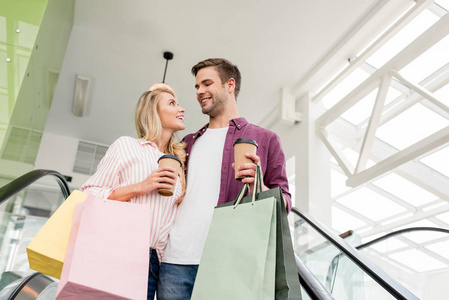 在购物中心的自动扶梯上用纸袋和咖啡杯购物的年轻夫妇的低角度视图