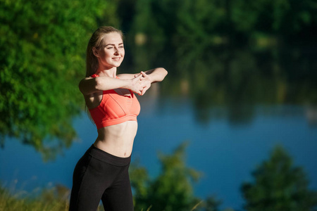 年轻的金发美女，在大自然中锻炼。运动女孩穿着红背心站在河边的公园