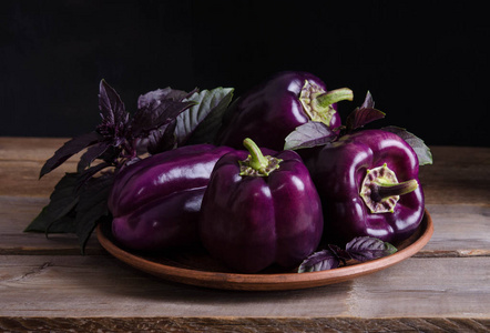 新鲜蔬菜。黑色背景上老土气木桌上的罗勒叶子上的深紫色辣椒