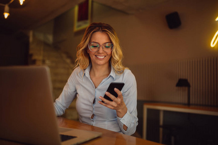 戴眼镜的年轻妇女正在检查智能手机上的新信息, 而坐在咖啡馆和工作的笔记本电脑