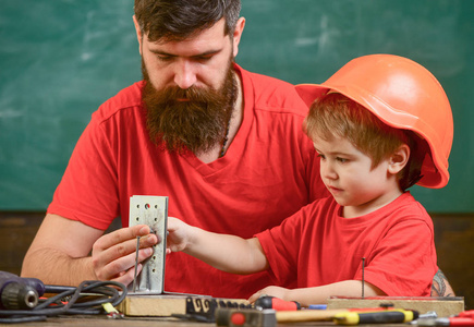 男孩, 孩子忙着戴着防护头盔做手工, 修理爸爸。小助手的概念。父亲用胡子教小儿子用工具在教室里, 黑板上的背景