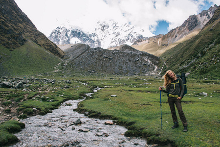 在 Salkantay 山徒步旅行期间, 带着旅行背包站在河边, 在相机上微笑的欢快年轻女子的全长肖像。女性徒步旅行者在流浪期间