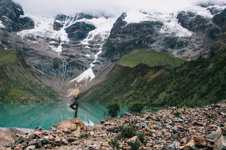 在高山岩石上做瑜伽的年轻女子旅行者的背观欣赏着壮观的湖泊和美丽的山脉, 覆盖着积雪和云层。在 Salkantay 的旅游平衡姿态