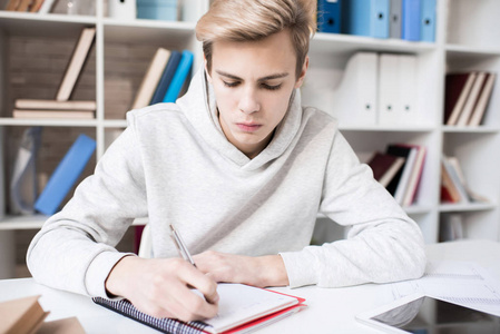 年轻集中的白种男性学生写笔记在抄写时, 当坐在书桌在教室里