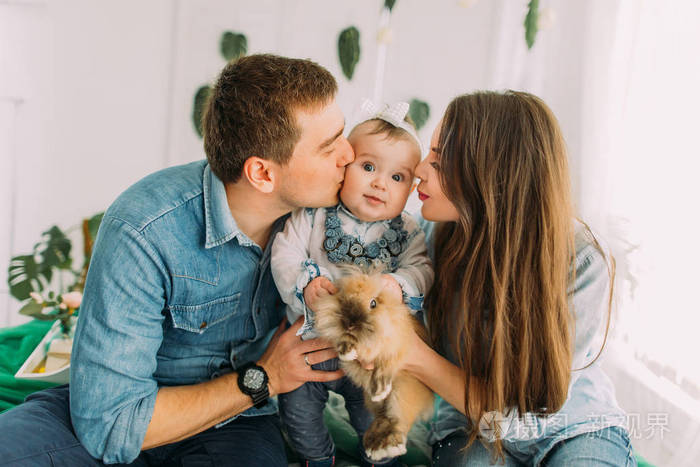 家庭肖像.父母亲把婴儿亲吻到脸颊, 抱着可爱的兔子.