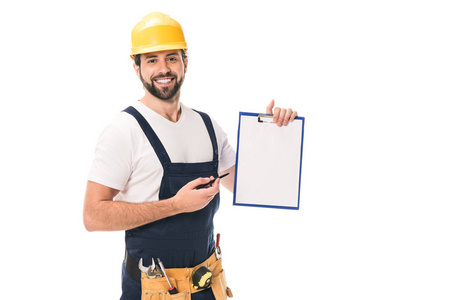 快乐建筑工人在硬帽子和工具皮带持有空白剪贴板和微笑着在白色的相机隔离