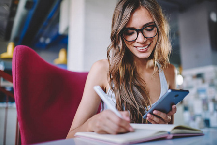 积极的年轻妇女在眼镜做笔记在记事本坐在桌子上, 并使用免费 wifi 连接智能手机的搜索信息。快乐的学生阅读通知