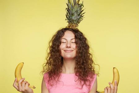 年轻女性用菠萝代替帽子, 保持眼睛紧闭, 手里拿着香蕉作为手印标志, 享受宁静的气氛, 与世隔绝的黄色演播室背景。人和冥想概念