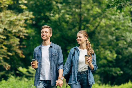 微笑的年轻夫妇与纸杯咖啡走在一起绿色公园