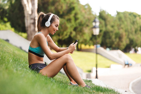 快乐的年轻运动员穿着运动装在耳机休息, 而坐在草地上的手机户外