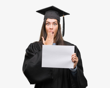 年轻的西班牙裔女子身穿毕业制服持有文凭纸捂着嘴用手震惊的错误, 表达恐惧, 害怕沉默, 秘密概念