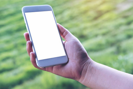 手持和使用白色智能手机, 户外和绿色自然背景下的空白屏幕的样机图像