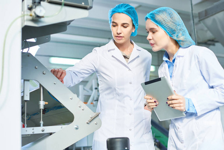 两名身穿实验室大衣的年轻女子在现代食品厂工作的肖像和微笑, 同时检查生产过程中的电源单位站在车间, 复制空间