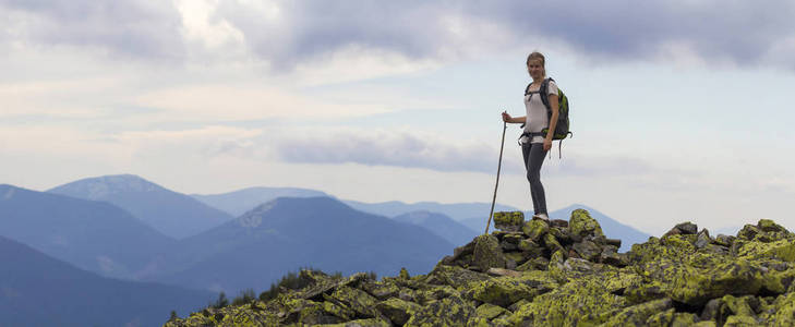 年轻苗条的金发旅游女孩与背包和棍子站在岩石顶部对明亮的蓝色早晨天空在雾山范围全景背景。旅游旅行和登山概念