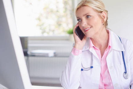 一名女医生坐在电脑前, 在与病人预约时使用手机的镜头
