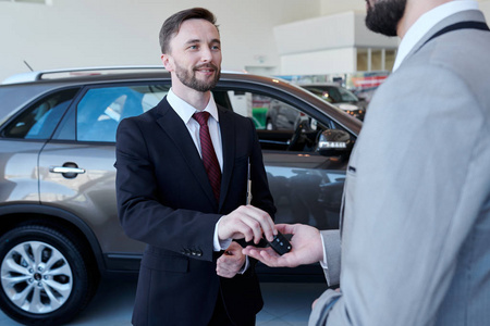 腰部向上的微笑英俊的汽车推销员给汽车钥匙客户站在豪华经销商展厅的纵向