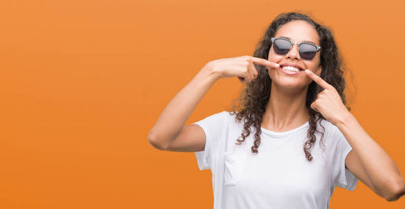 年轻的西班牙裔妇女戴着太阳镜微笑着自信地显示和指着手指的牙齿和嘴巴。健康理念