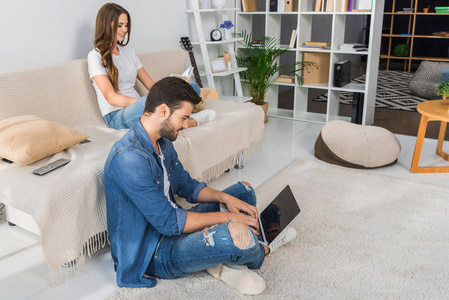微笑的人使用膝上型电脑和坐在地板上, 而他的女朋友在沙发上看书在家里的侧面视图