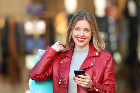 快乐的购物者摆在街上拿着一个智能手机和购物袋的背景下的店面