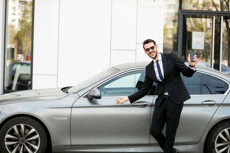 超级司机穿着优雅的西装在一辆豪华轿车前展示他的手机, 外面的街道上的办公室大楼前